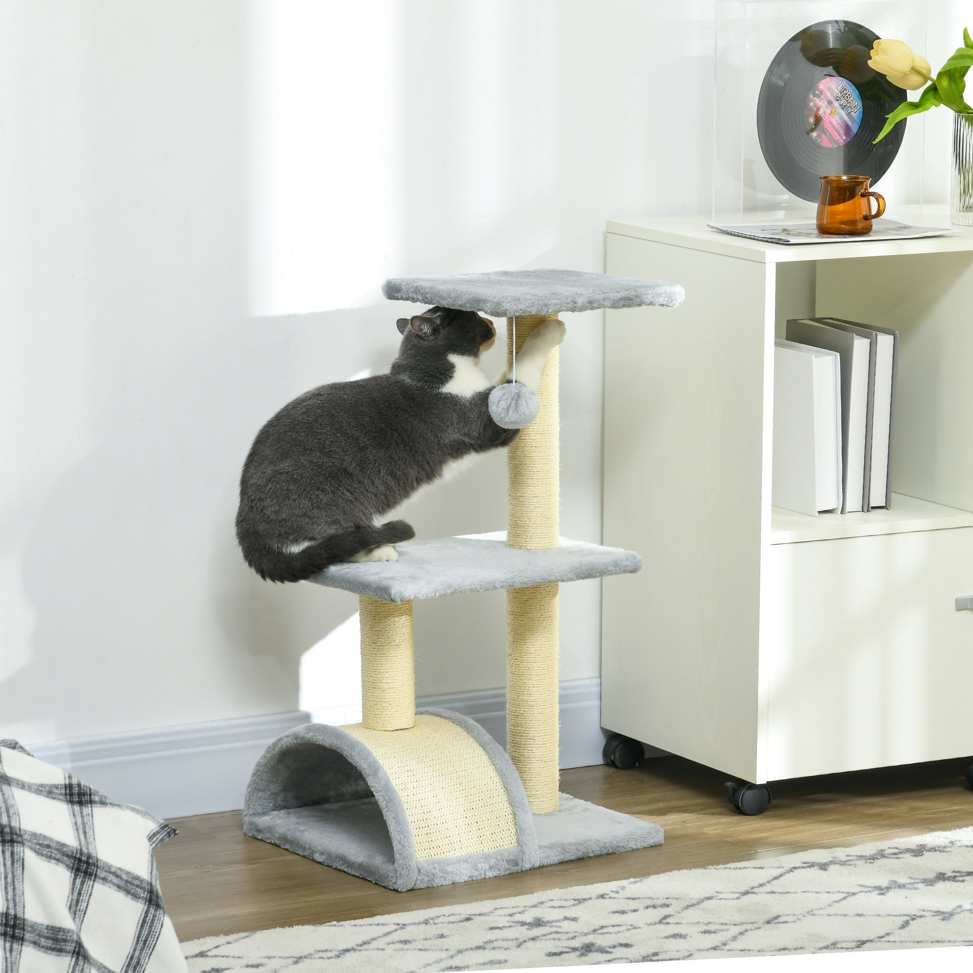 En katt som klöser på ett stilfullt ljusgrått och cremévitt klösträd placerat i ett vardagsrum, utrustat med en böjd klösbräda och en mjuk utsiktsplats.