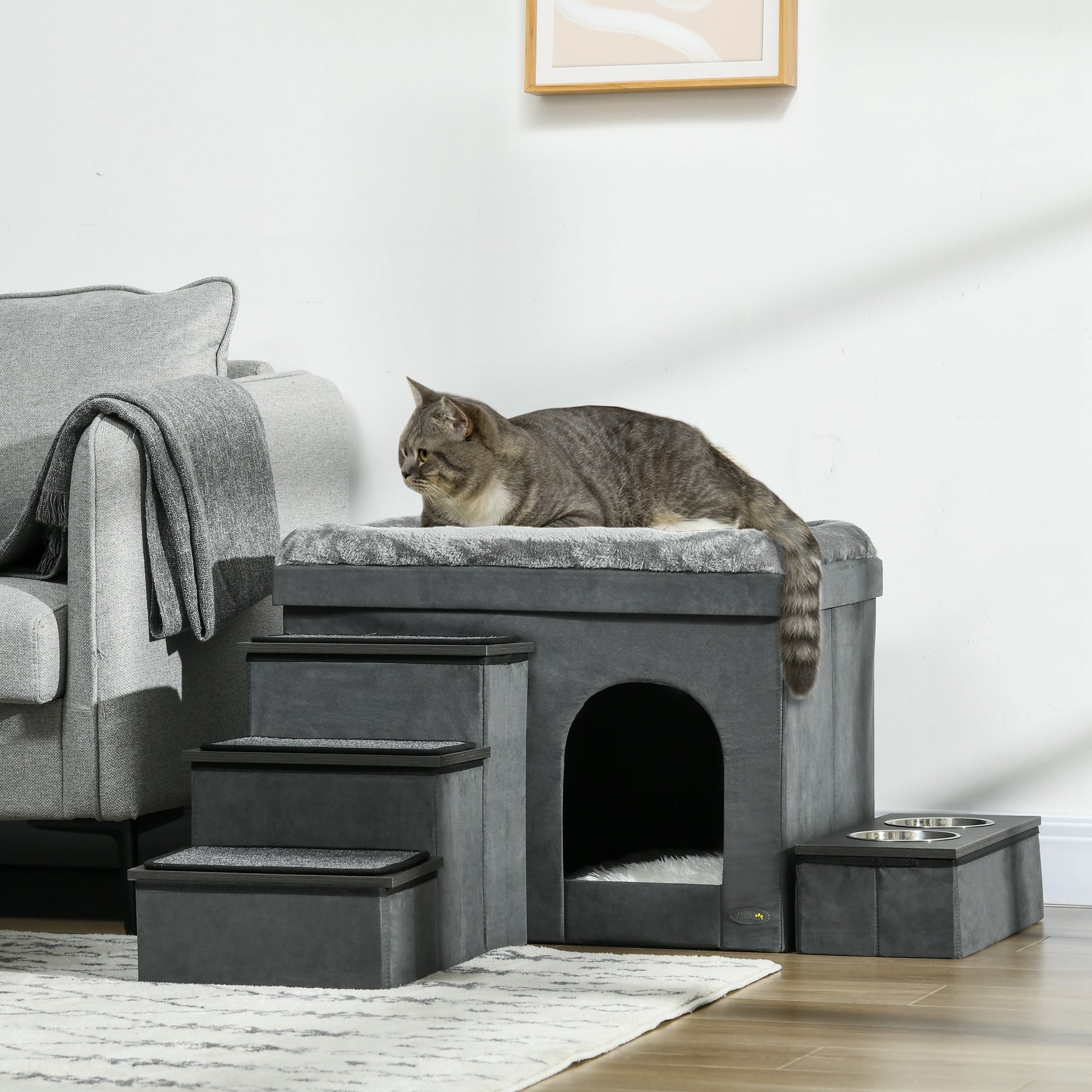 En katt vilar på toppen av en mångsidig grå husdjursmöbel med trappsteg, inbyggt hus och matstation, placerad i ett vardagsrum bredvid en fåtölj.