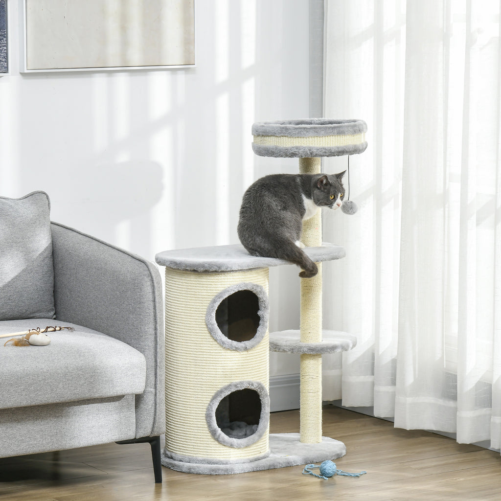 En katt sitter på en plattform på ett mångsidigt grått klösträd utrustat med en mysig dubbelgrotta och sisalpelare, placerat bredvid en grå soffa.